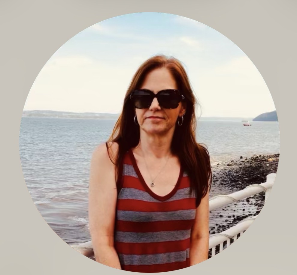 A woman with long brown hair wearing sunglasses and a striped tank top, standing by the water with a scenic view in the background.