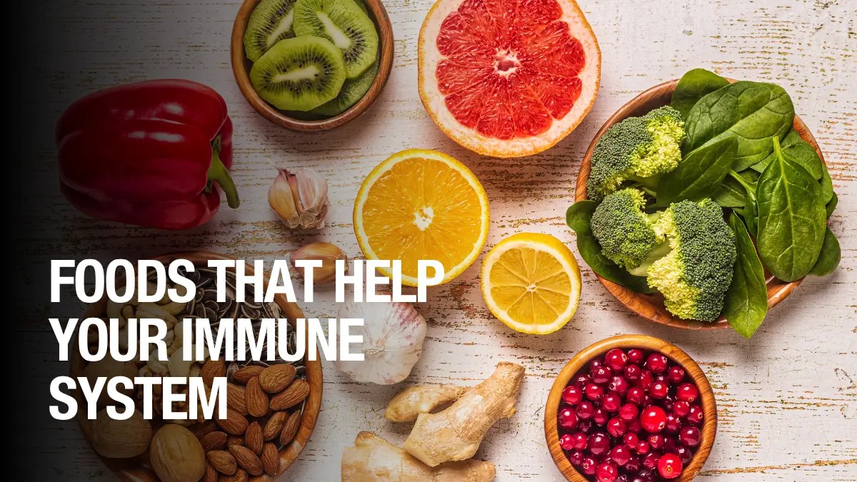 Flat lay of healthy foods including broccoli, spinach, grapefruit, orange, lemon, kiwi, garlic, red pepper, almonds, ginger, and cranberries. Text overlay reads: “Foods That Help Your Immune System.”
