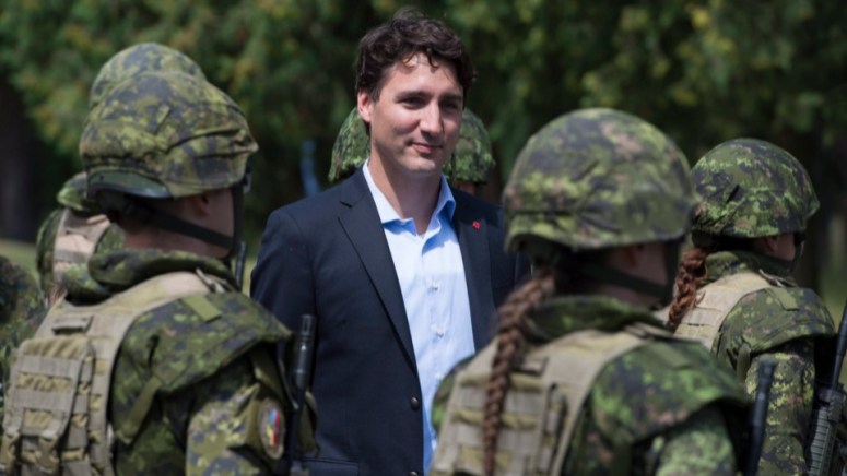 Un homme en costume avec un t-shirt bleu s'approche d'un groupe de soldats canadiens en uniforme militaire, lors d'un événement en extérieur.