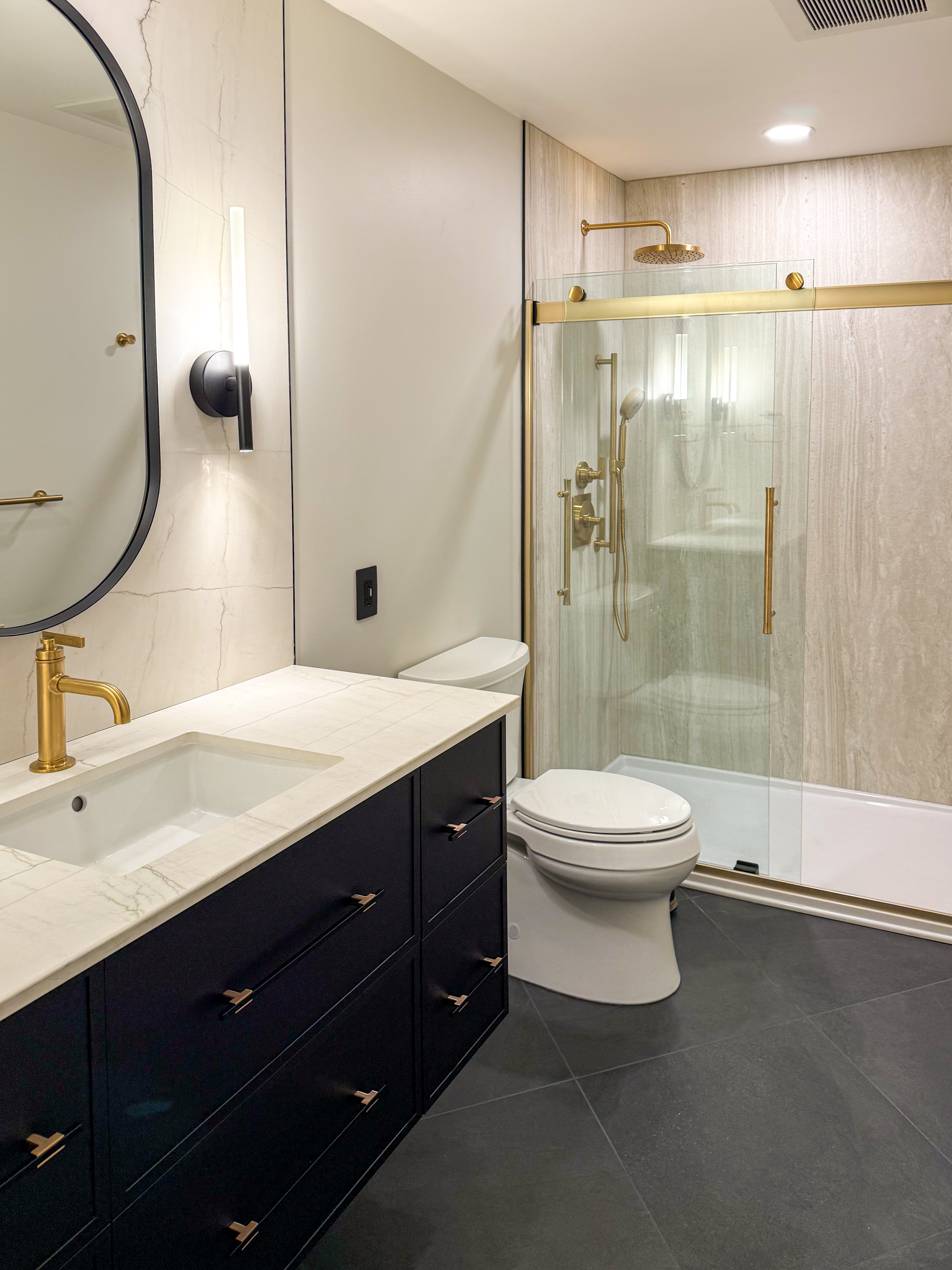 Montblanc River Spa Bathroom with river-washed porcelain shower wall panels, stone resin shower base, Kohler gold shower fixtures, sliding glass shower door, slate-look floor tile, floating satin black vanity, Montblanc porcelain vanity top, black sconces, and gold faucet.