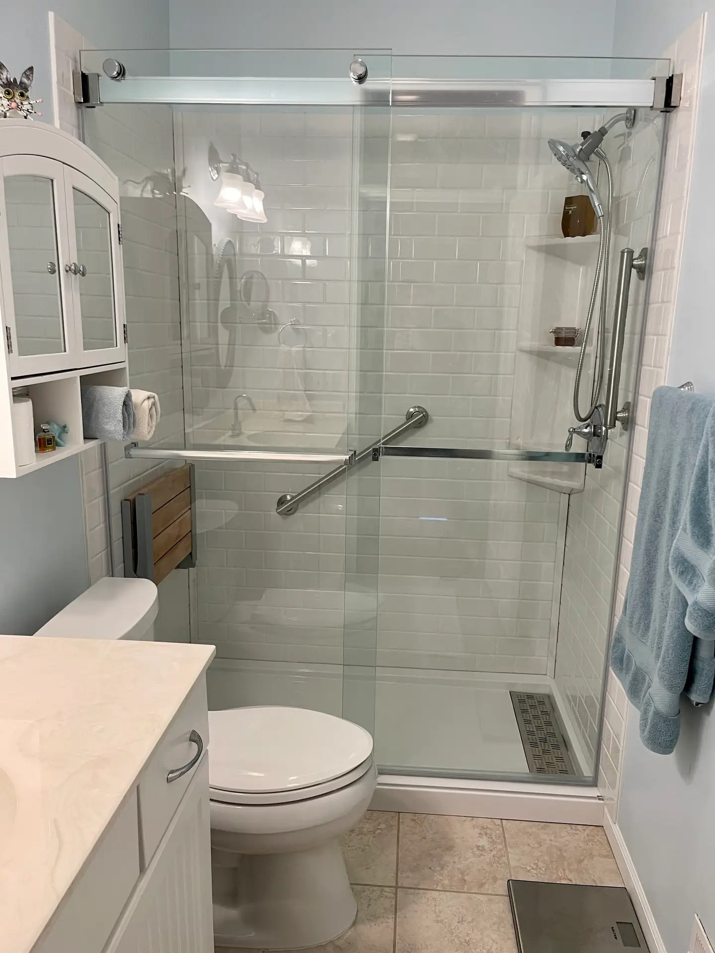 Teak Comfort Subway Shower with white subway tile pattern stone resin walls, stone resin shower base, sliding glass door, corner caddy shelf, handheld shower, grab bars and teak wood fold down bench.