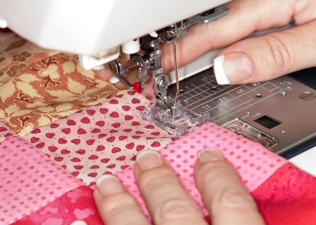 A close-up of an individual with manicured fingers using a white sewing machine to stitch a red and pink quilt. A quick guide to adding texture to your quilts will help you create better designs.