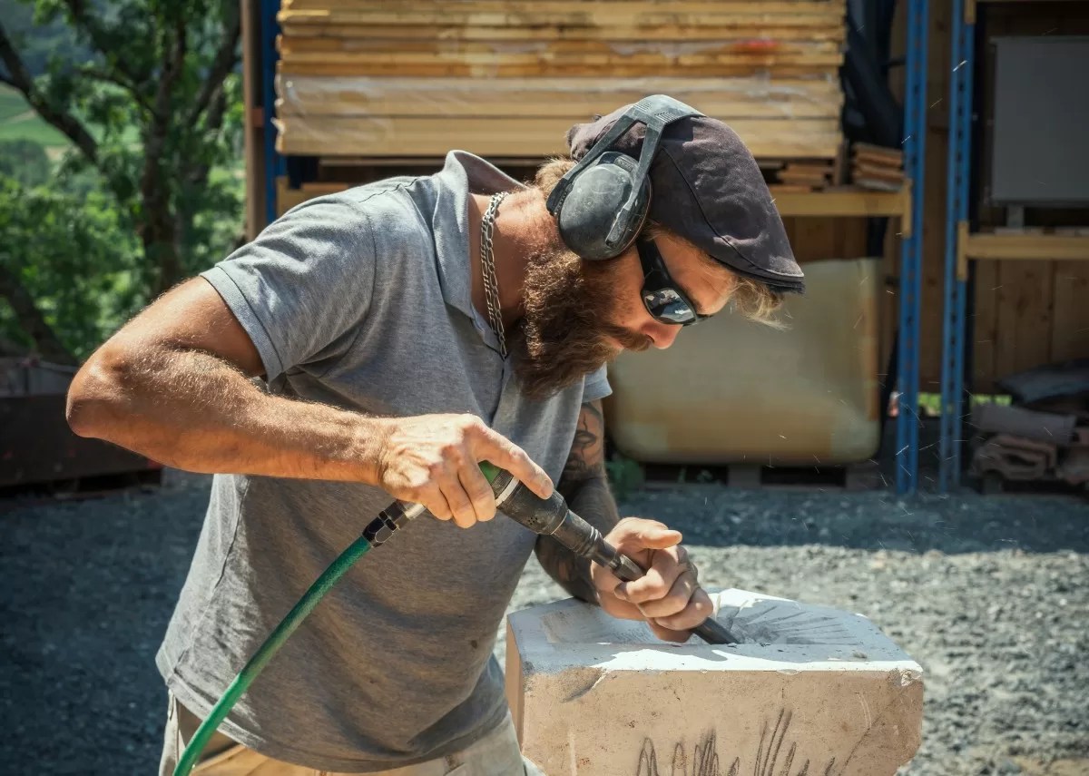 A stone sculpter using a pneumatic chisel to carve a design into a block of light gray stone outside.