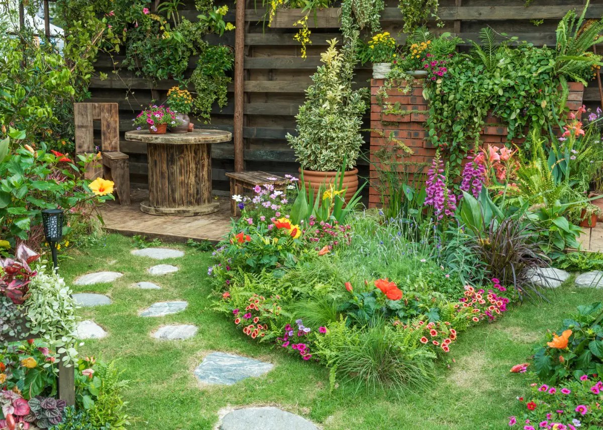 A lush garden with colorful flowers, stepping stones, potted plants, and a rustic wooden table and chair.