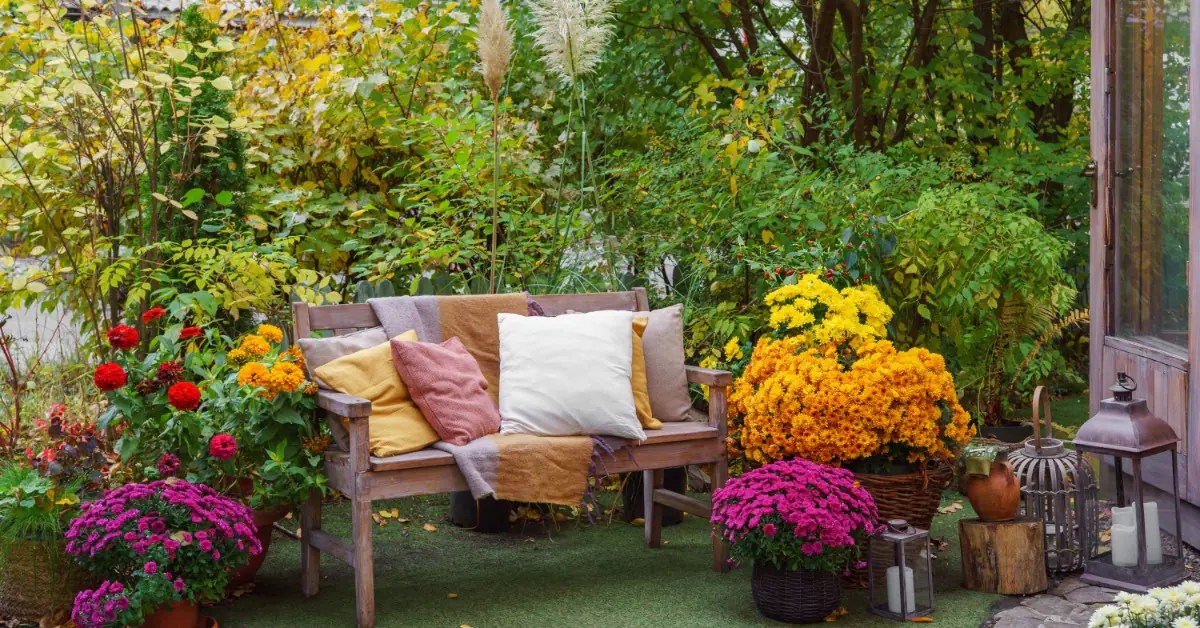 A wooden garden bench with colorful pillows surrounded by bright flowers, greenery, and decorative lanterns.