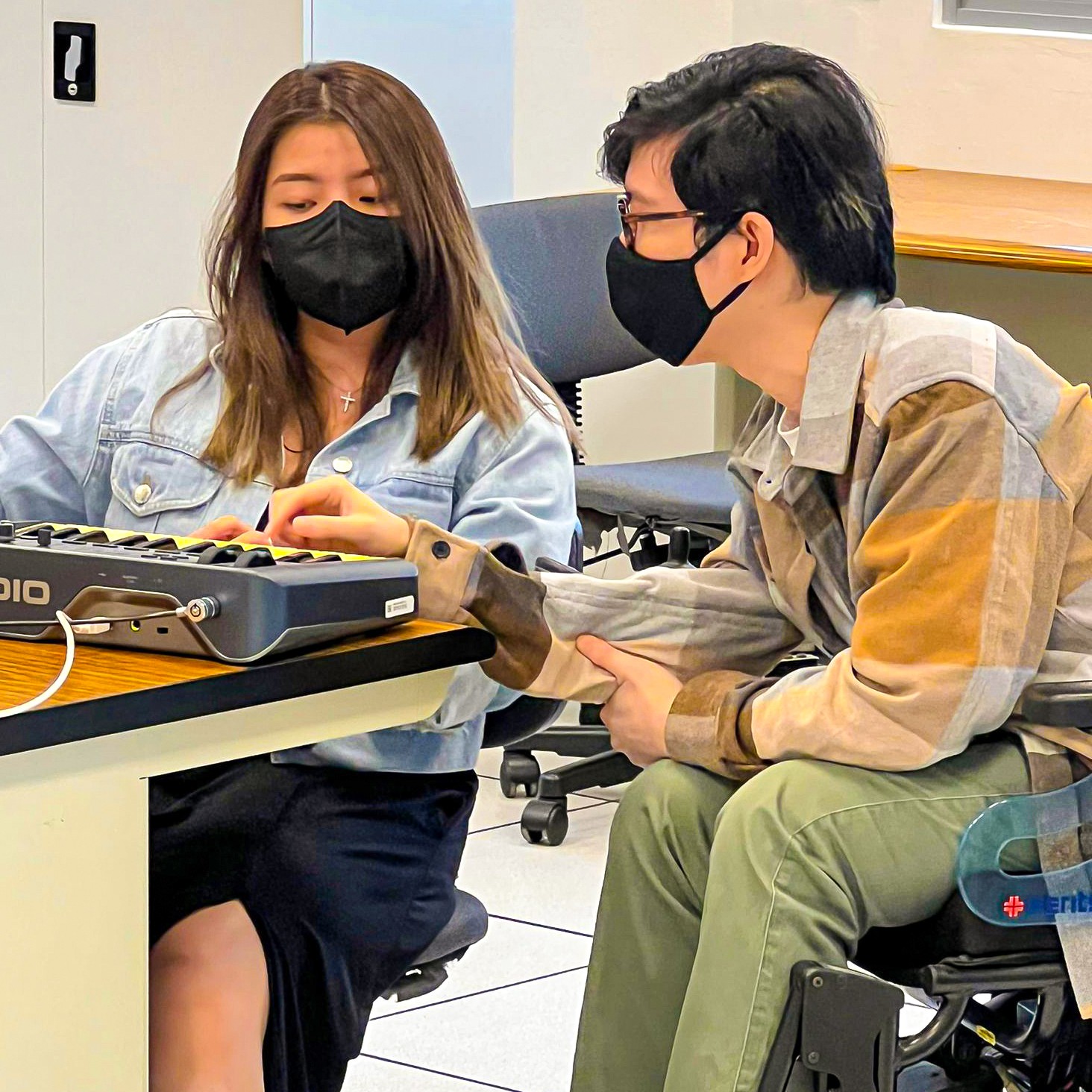 This image features a young wheelchair user providing guidance to a participant on how to create music with an electronic keyboard.