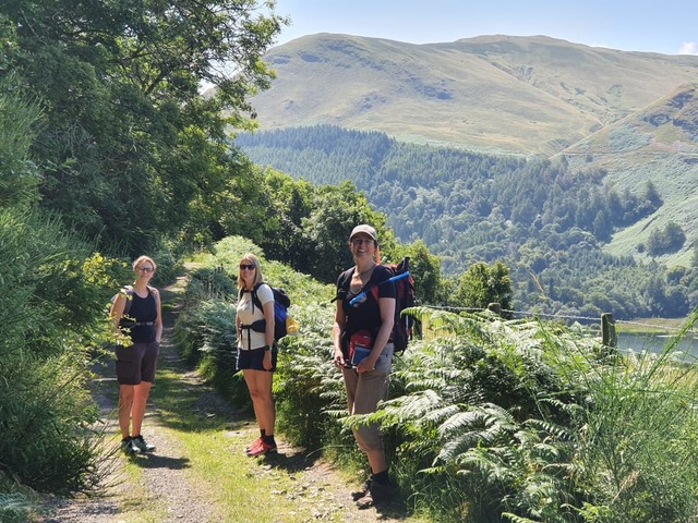 3 women stand on a rural path with ferns in verges and fells in distance.