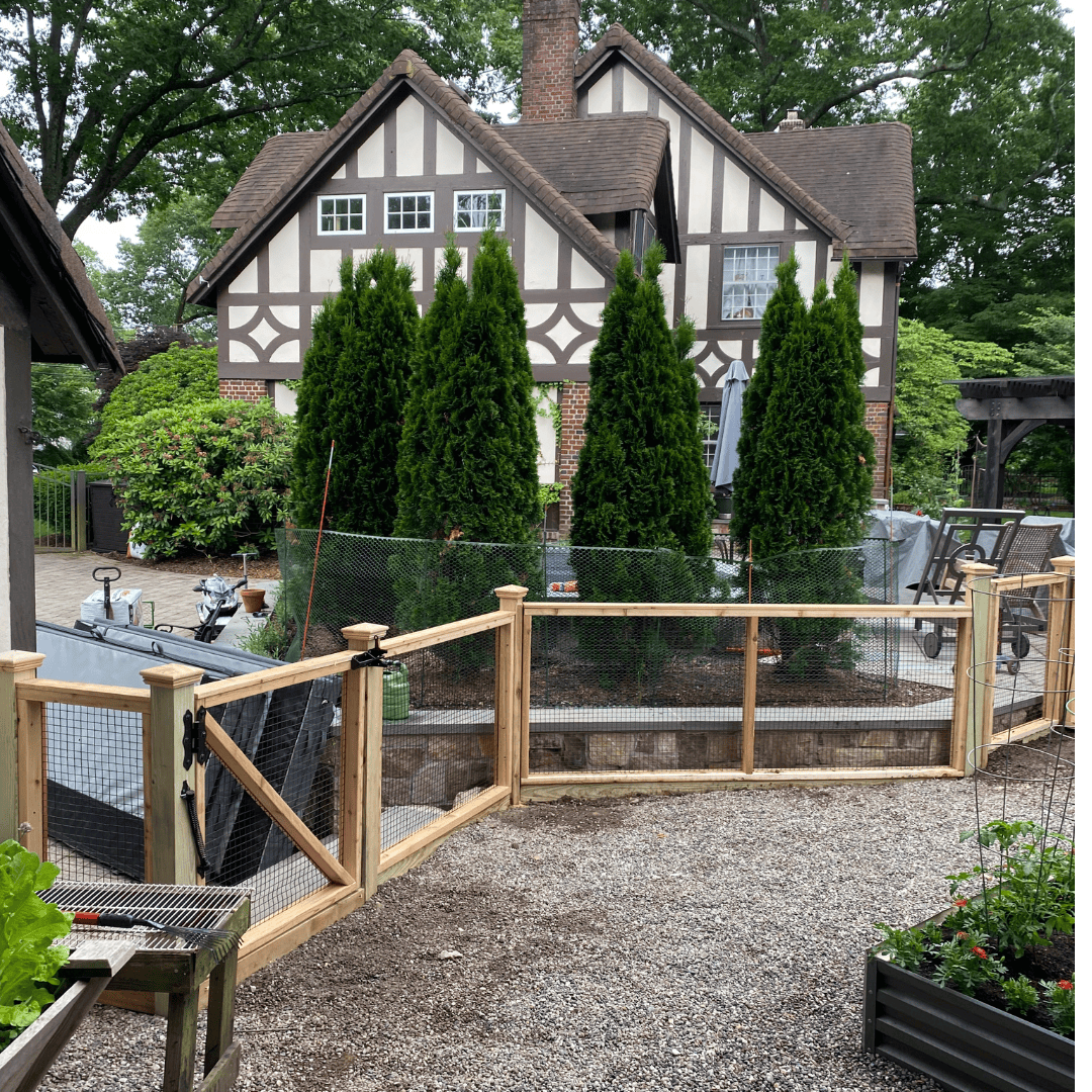 Wood and wire garden fencing around a vegetable garden with a tudor style home in the background