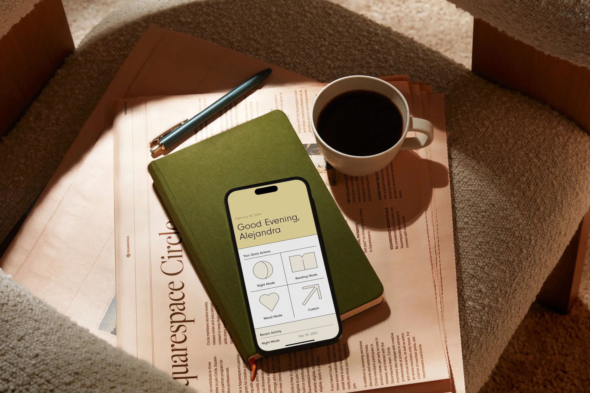 A green notebook with a smartphone on top, a cup of black coffee, a pen, and newspaper on a beige chair and carpet.