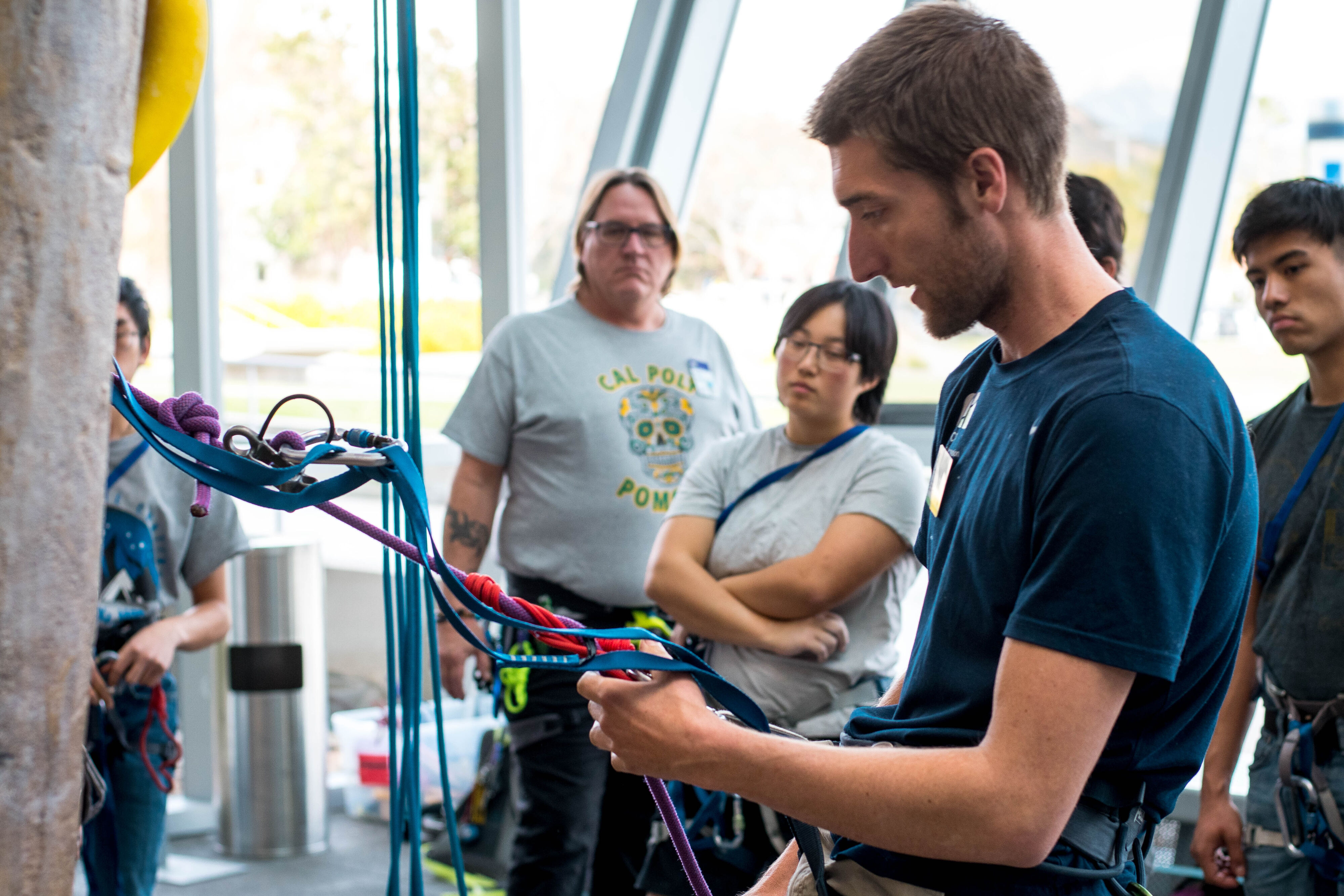  A man in climbing gear is demonstrating rope techniques to a group of attentive onlookers inside a climbing gym.
