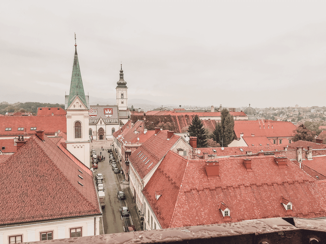 Overlooking the terracotta rooftops of Zagreb, Croatia, this image captures the city's old-world charm with its historic buildings and church spires, nestled under a hazy sky
