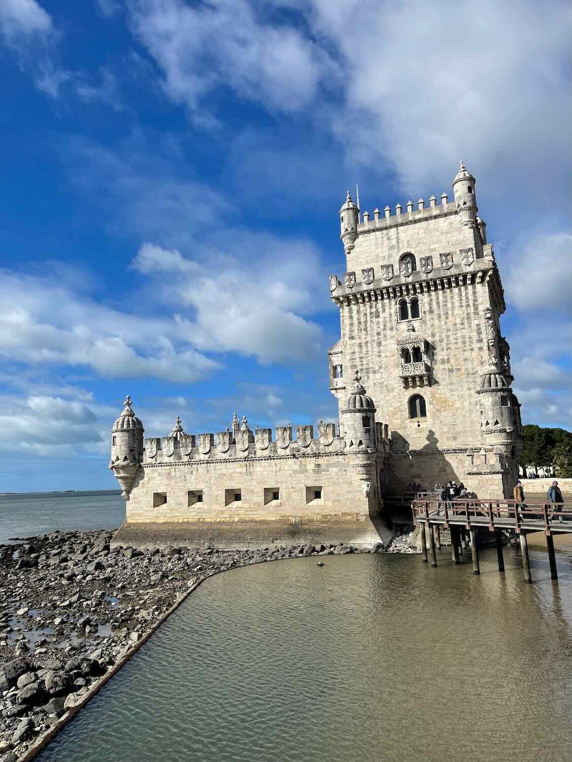 A close-up view of the iconic Bel&eacute;m Tower in Lisbon, standing majestically by the water. The stone fortress features intricate details and turrets, with a bright blue sky and scattered clouds in the background.