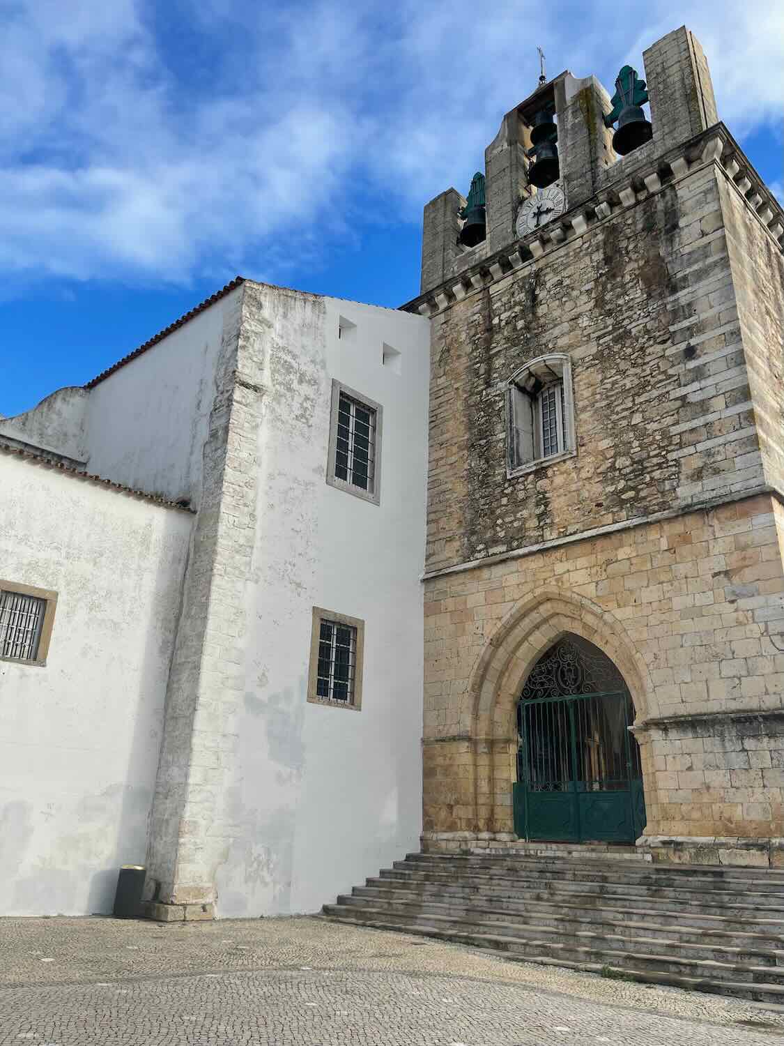 A medieval stone church with a Gothic arched doorway and a prominent bell tower rises against a bright blue sky in Faro, Portugal, showcasing the city's rich architectural heritage.