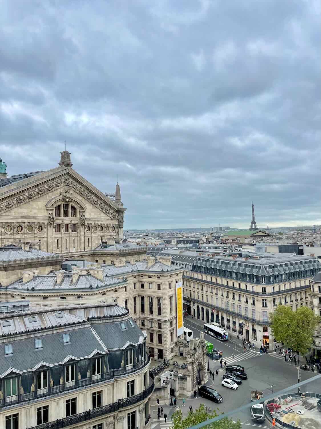 elevated view of Paris from a rooftop, showcasing the ornate architecture of the Opera Garnier, with the iconic Eiffel Tower visible in the distance under a cloudy sky.