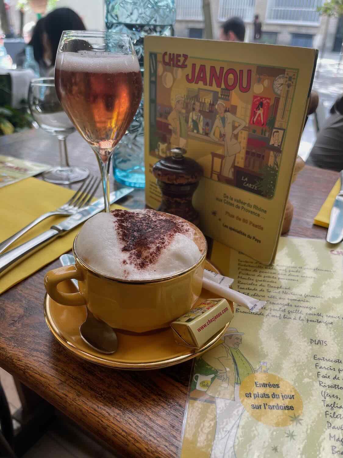 A table setting at Chez Janou featuring a cappuccino with a heart-shaped design in the foam and a glass of ros&eacute;, accompanied by a colorful menu, creating a cozy and welcoming dining experience.