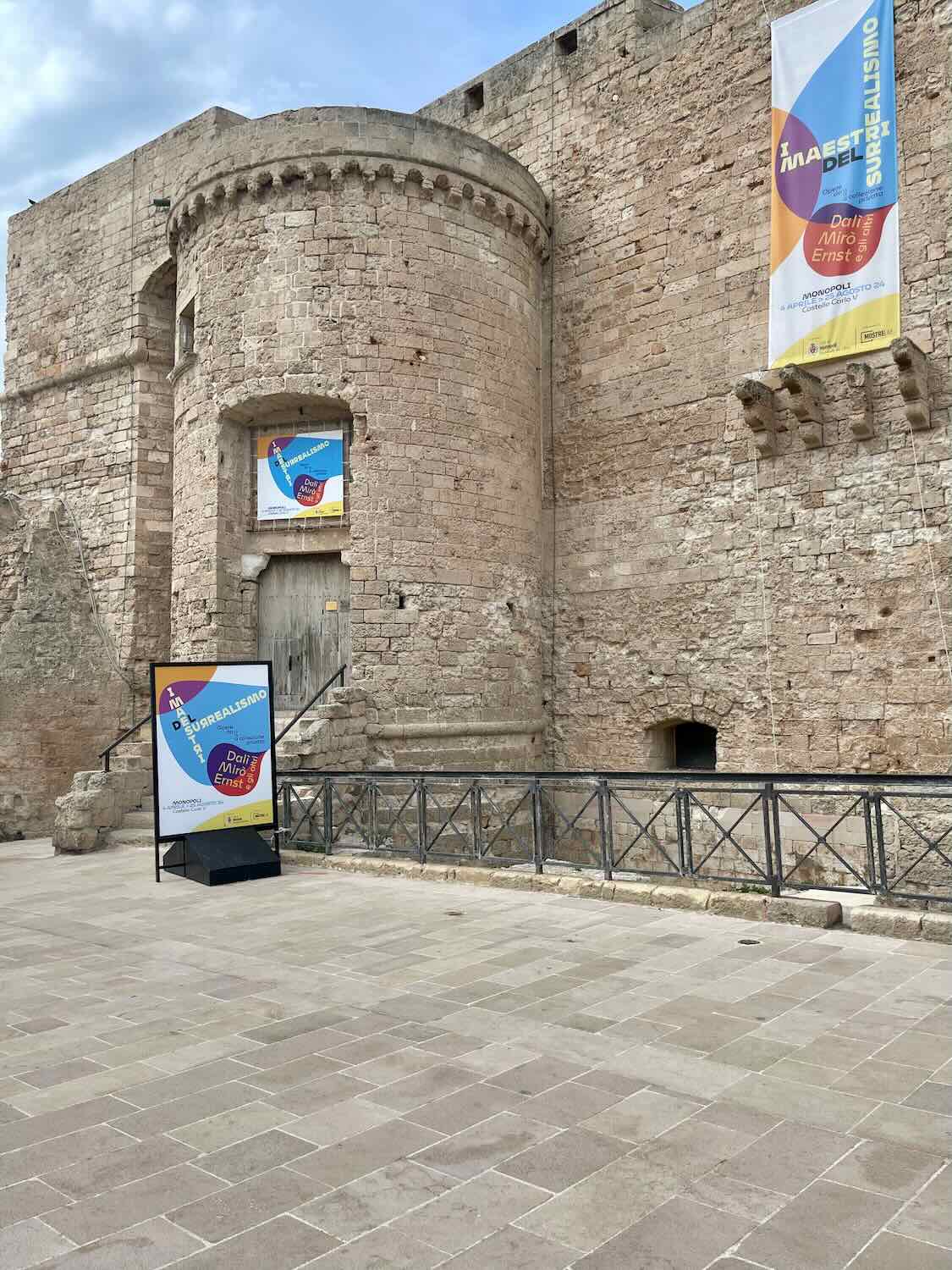 A view of an old castle with a circular tower and banners promoting an art exhibition. The castle is made of light-colored stone, and the ground is paved with large stone tiles.