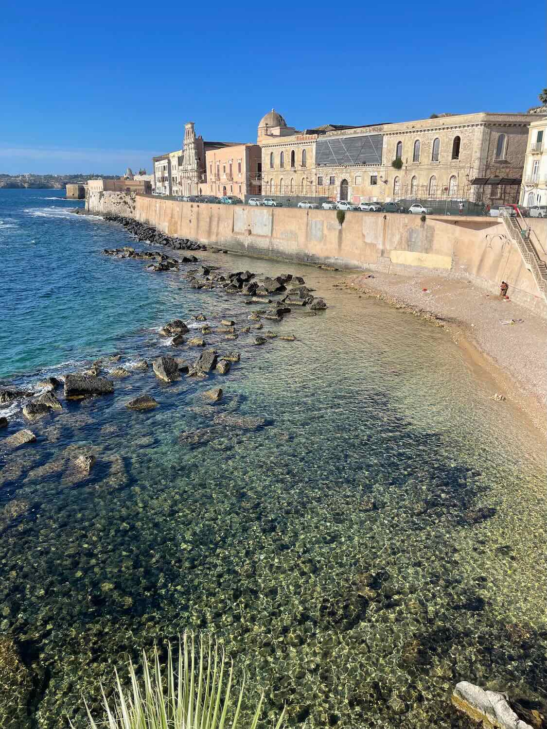 A scenic coastal view with clear blue water and rocky shoreline. In the background, there are historic buildings along a waterfront promenade under a clear blue sky.