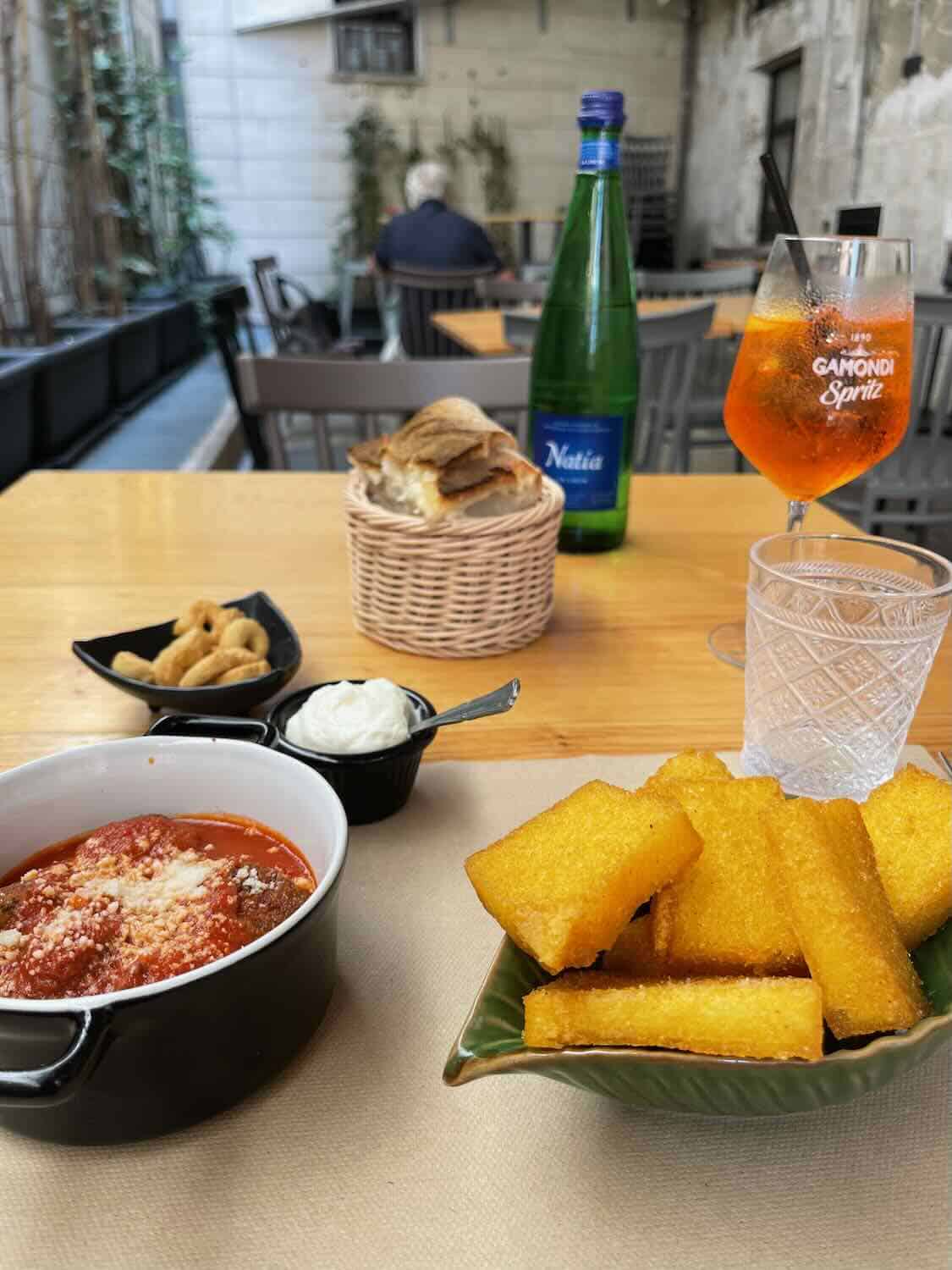 A table set with an assortment of Italian foods, including a dish of meatballs in tomato sauce, a plate of fried polenta sticks, a small bowl of cream, and a basket of bread. A glass of Aperol Spritz and a bottle of sparkling water are also on the table, with an outdoor caf&eacute; setting in the background.