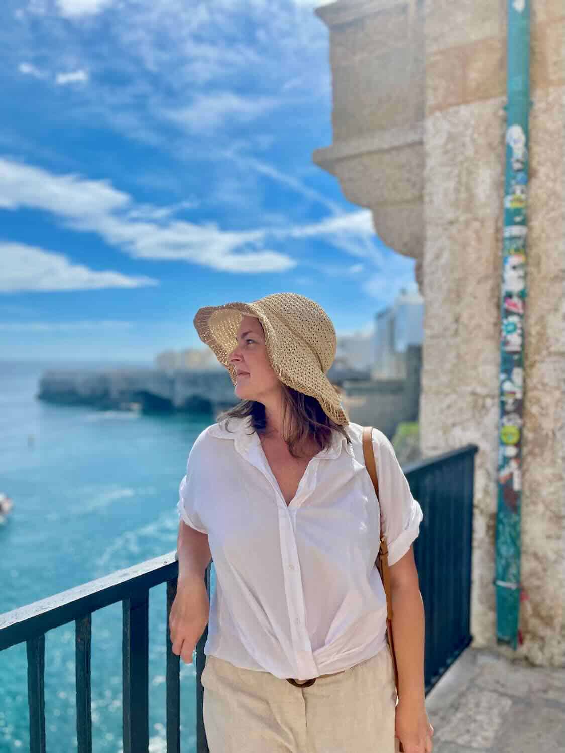 A woman wearing a white shirt and beige skirt with a wide-brimmed hat, standing by a railing overlooking the clear blue sea in Polignano a Mare. The background includes historic buildings and a partly cloudy sky.