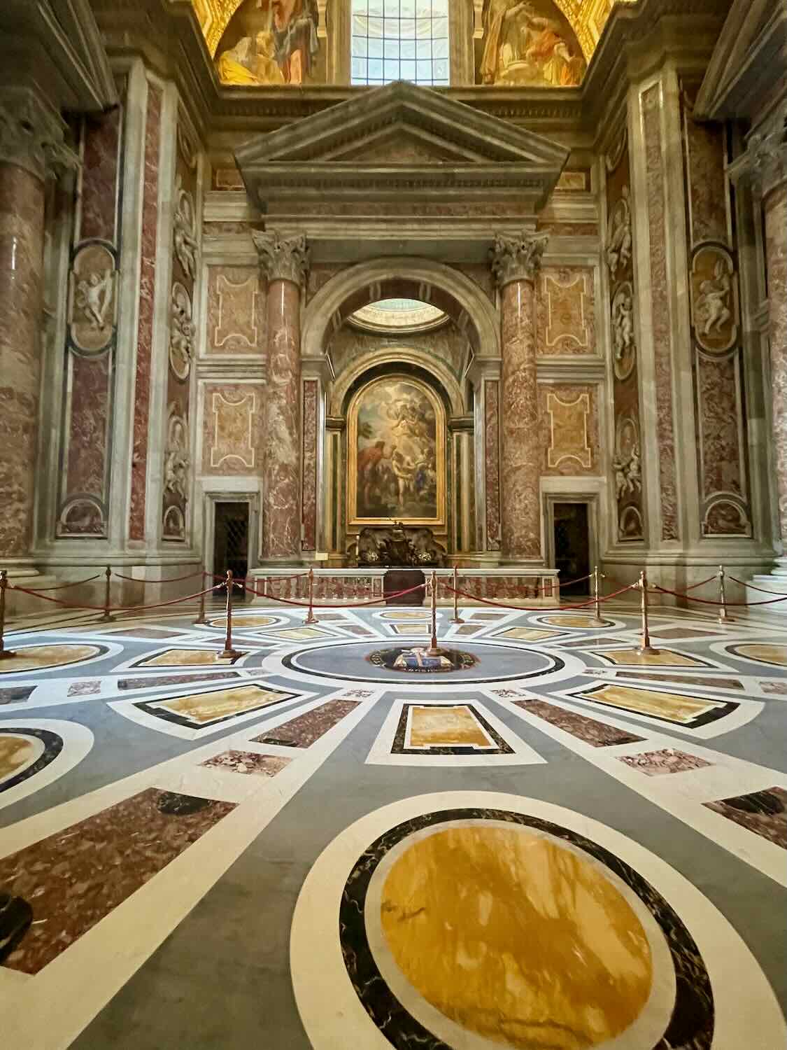 An interior view of a grand room in the Vatican, showcasing an intricate marble floor with circular patterns, tall marble columns, and an ornate altar with religious artwork and statues.

