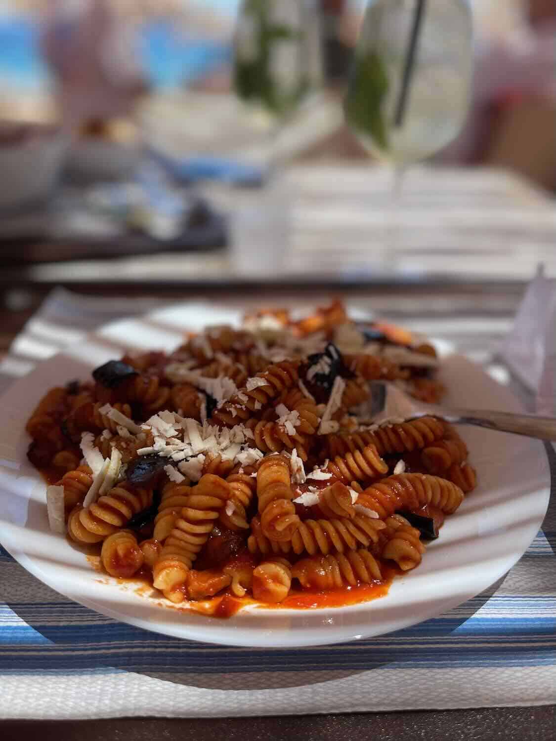 A close-up view of a plate of pasta with tomato sauce and grated cheese. The background is slightly blurred, showing a beachside dining area with drinks and a blue sea.