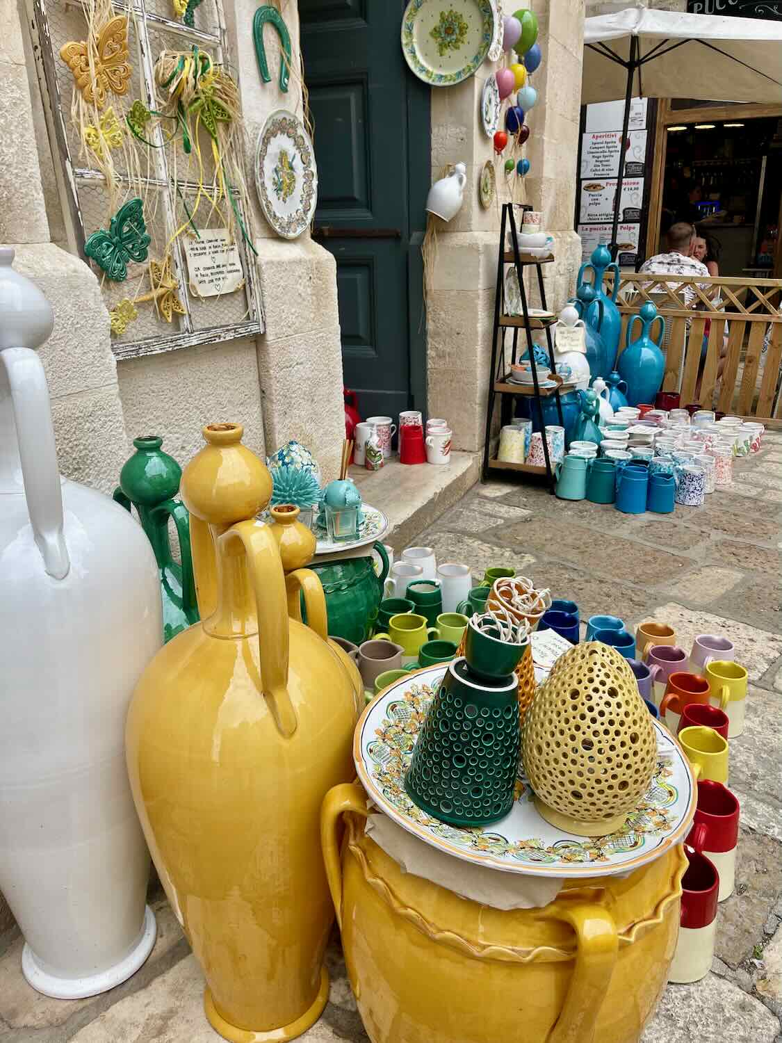 A colorful display of ceramic pottery outside a shop in Polignano a Mare. The assortment includes large yellow and white vases, mugs, plates, and decorative items arranged on shelves and tables.