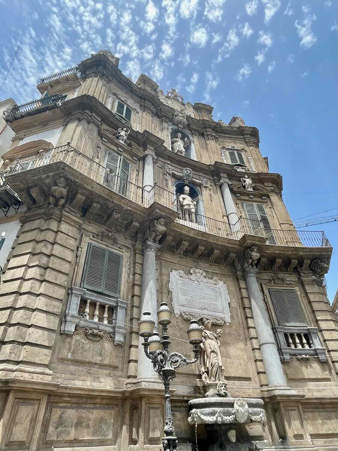 The intricate fa&ccedil;ade of Quattro Canti, a historic Baroque square in Palermo, Sicily, featuring statues and detailed carvings. The building's weathered stonework stands out against a partly cloudy sky.