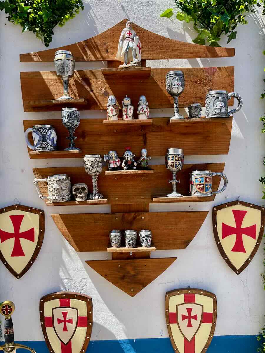 A wooden wall display in &Oacute;bidos, adorned with medieval-style goblets, figurines of knights, and shields bearing the red cross of the Knights Templar. This display gives a glimpse into the medieval culture of the town.