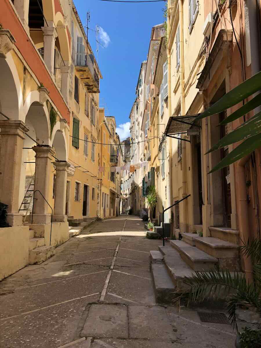 A charming alley in Corfu, Greece, showcasing arched buildings and pastel-toned facades under a vivid blue sky. Laundry hangs between the buildings, adding to the lively, authentic Mediterranean ambiance.