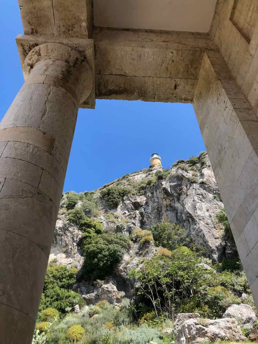 A view through stone columns reveals a rugged cliff adorned with lush greenery in Corfu. At the top, a lighthouse stands against the vibrant blue sky, adding to the dramatic scene.