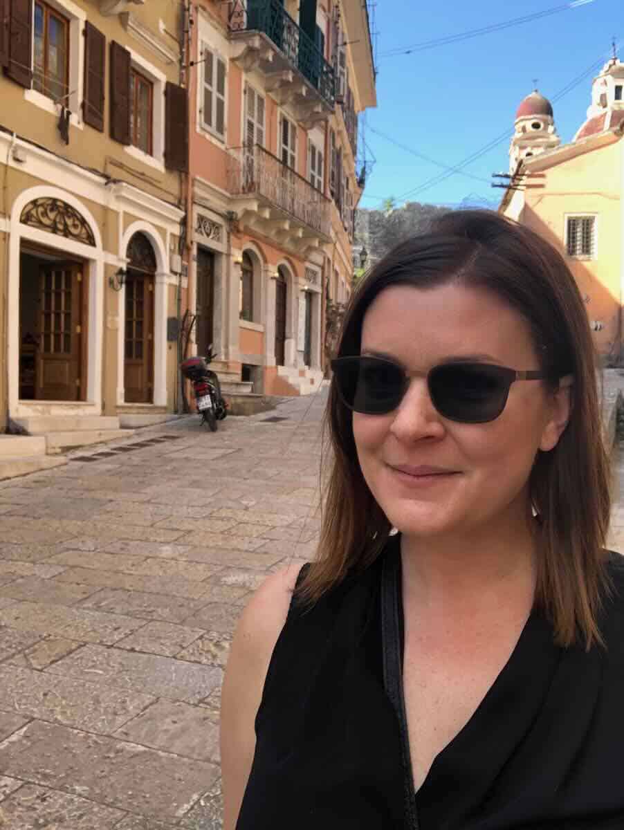 Melissa wearing sunglasses and a black top smiles while standing on a stone-paved street in Corfu, Greece. The background features pastel-colored buildings with balconies, a parked scooter, and a church dome visible in the distance.