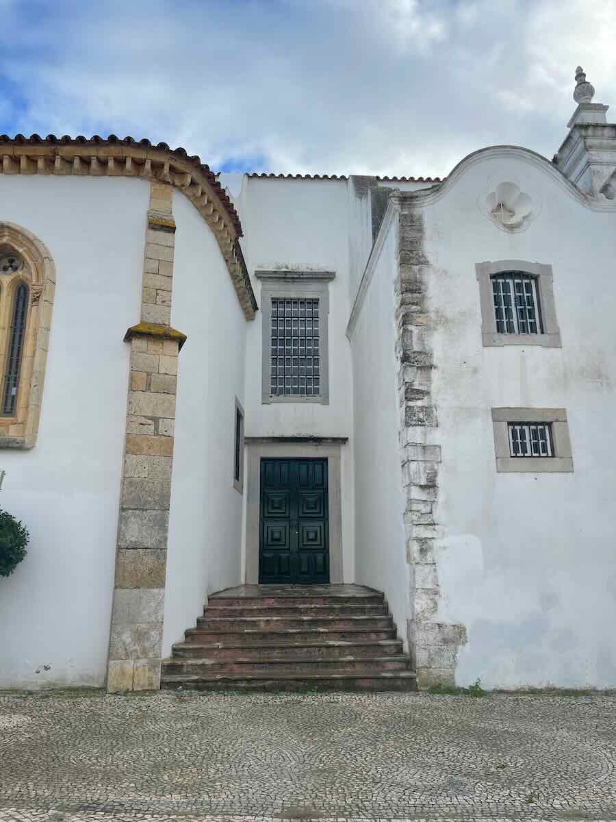 A historic whitewashed building in Faro, Portugal, featuring a mix of architectural styles, arched windows, and a green wooden door at the top of a small stone staircase, set under a cloudy sky.