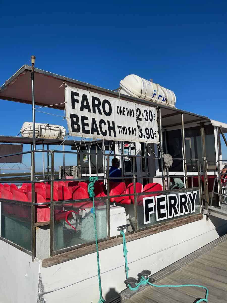 Ferry at Faro Beach with a sign displaying ticket prices: one-way for &euro;2.30 and round-trip for &euro;3.90, against a clear blue sky.
