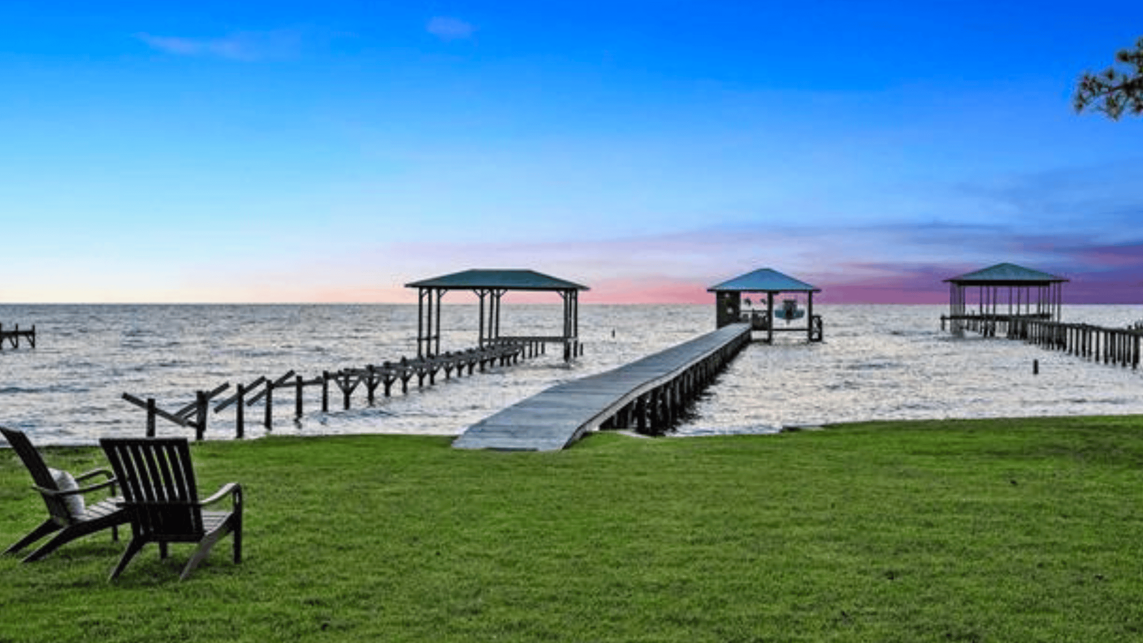 Waterfront lawn with Adirondack chairs facing a long private pier and covered boat houses on Mobile Bay at sunset in Fairhope, Alabama.