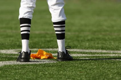 A referee stands on a football field with a yellow penalty flag on the ground next to his feet, ironically watching as the game's star player strikes a Heisman pose.