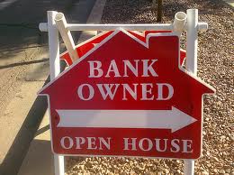 Red sign in the shape of a house with the text "BANK OWNED" and "OPEN HOUSE," along with a white arrow pointing to the right. Often seen after a strategic default, the sign is placed on a white stand outdoors.