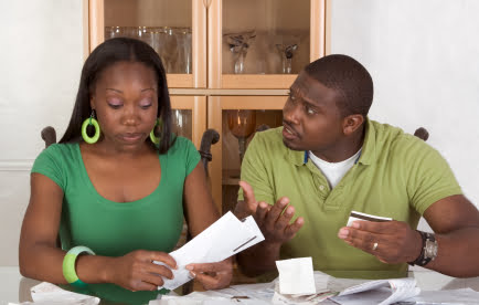 5 Ways To Spot a Debt Relief Scam 13 A woman and a man sit at a table covered with papers and envelopes, appearing to discuss or review the documents. The man looks concerned and gestures with his hands while the woman examines a piece of paper, possibly considering options for debt relief.