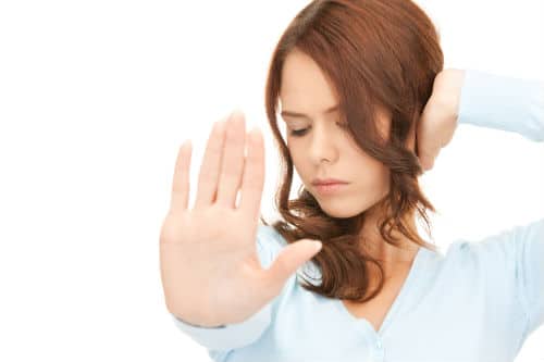 A millennial woman with brown hair and a serious expression extends her hand forward, palm facing out, while her other hand touches the side of her head. She is wearing a light blue shirt against a white background.