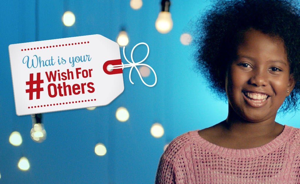 A smiling child stands beside a white tag sign that reads, "What is your #WishForOthers," with a gift card placed thoughtfully at its base, against a backdrop of hanging light bulbs.