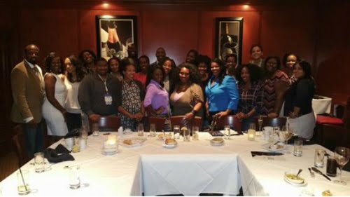 A group of bloggers of color posing together in a dimly lit room, standing around a table with empty plates and glasses.