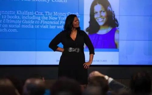 A woman stands confidently on stage, in front of a projection of her photo and a text presentation, showcasing her expertise as a financial planner.
