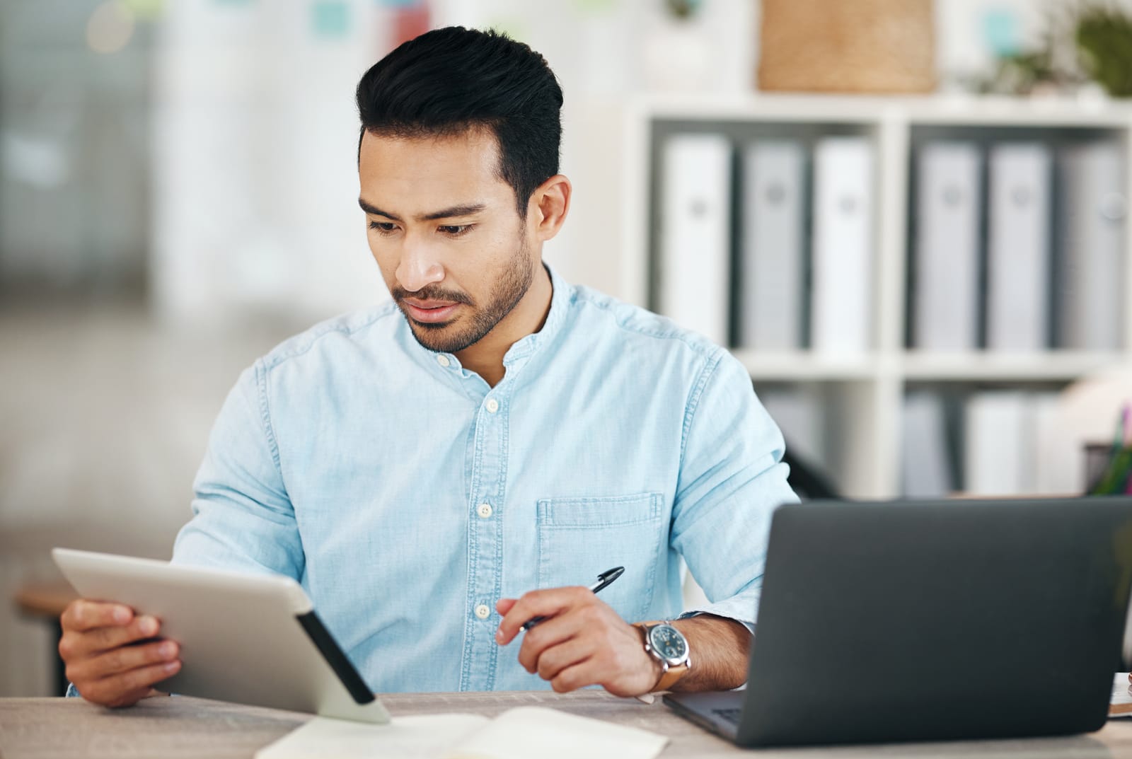 A man in a blue shirt sits at a desk using a tablet, with a laptop open beside him, acting as his loyal financial planner to help him stay on budget.
