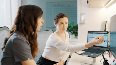 Two people sit at a desk in an office. One person, using AI-assisted tools, points at a laptop screen while the other looks attentively. team underperformance