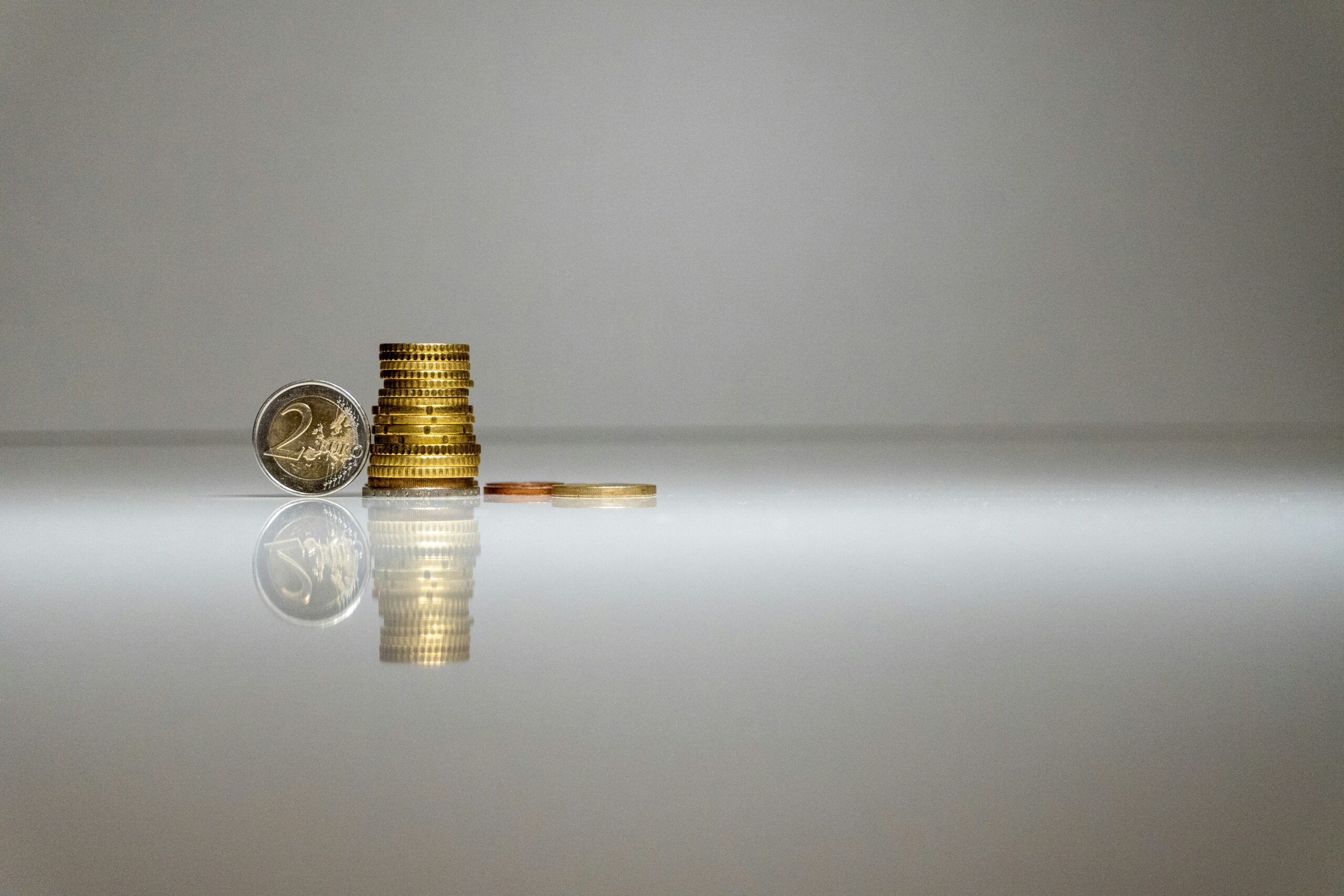 A stack of euro coins and a single two-euro coin stand on a reflective white surface against a plain background, symbolizing the start of your personal financial plan.
