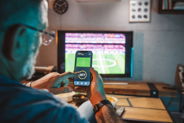 Mature men sitting on sofa at home and watching soccer game. He is using smart phone for sports betting .