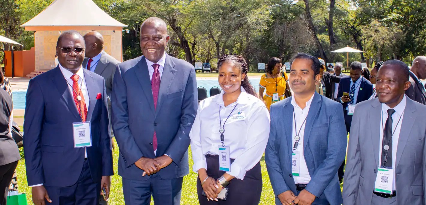 Five professionals in suits with lanyard badges pose for a photo at an outdoor conference