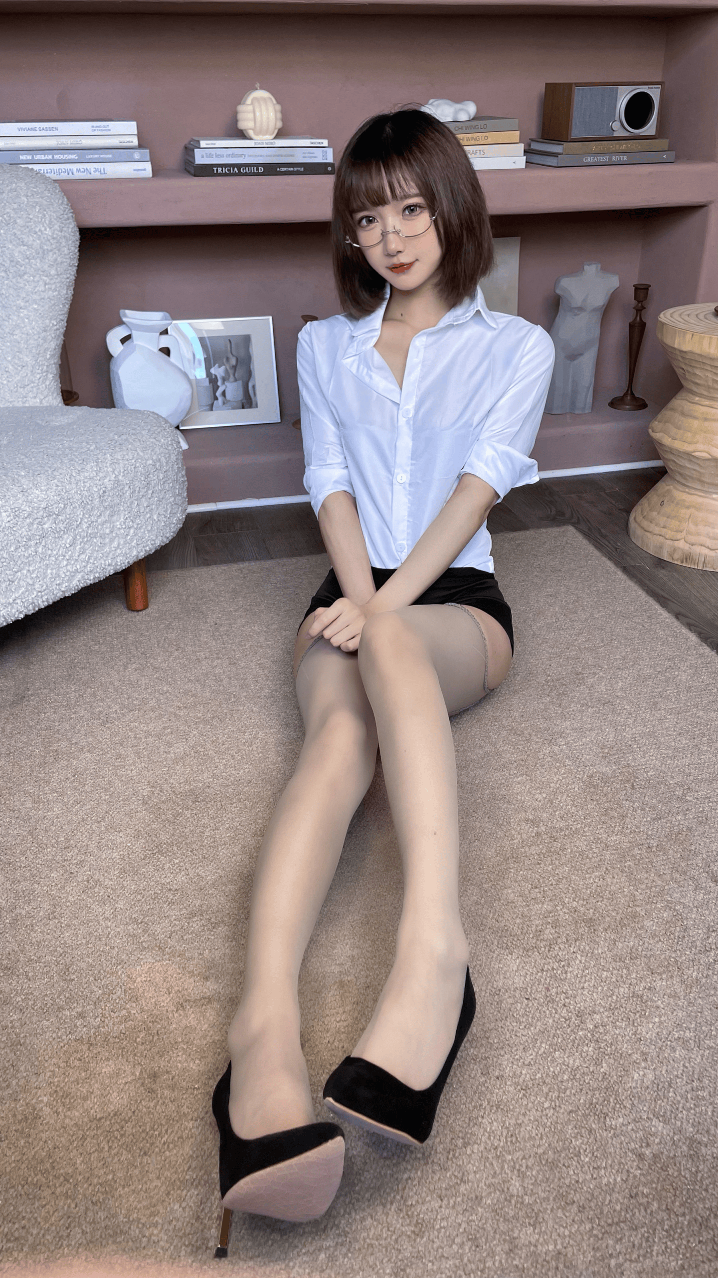 A young woman with short brown hair and glasses sits on a beige carpet wearing a white button-up shirt, black shorts, sheer stockings, and black high heels. She is positioned in a stylish room with mauve walls, built-in shelves displaying books and decorative objects, a gray textured chair, and wooden furniture.