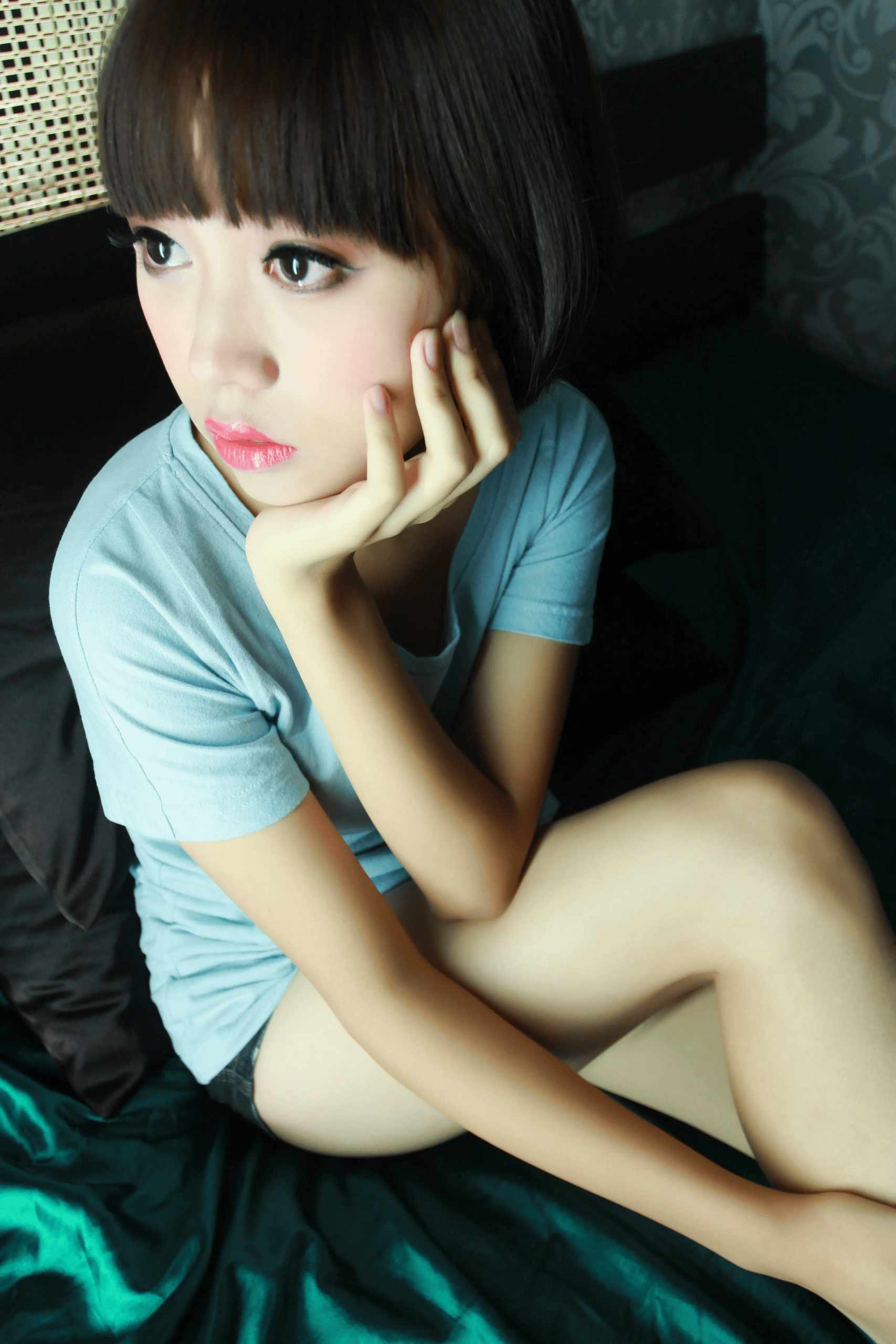 Portrait of a young East Asian woman with short dark bob cut and blunt bangs, wearing a light blue casual short-sleeve T-shirt, sitting on dark emerald satin bed sheets. She rests her cheek on one hand, gazes away to the side, and wears soft makeup with glossy bright pink lipstick.