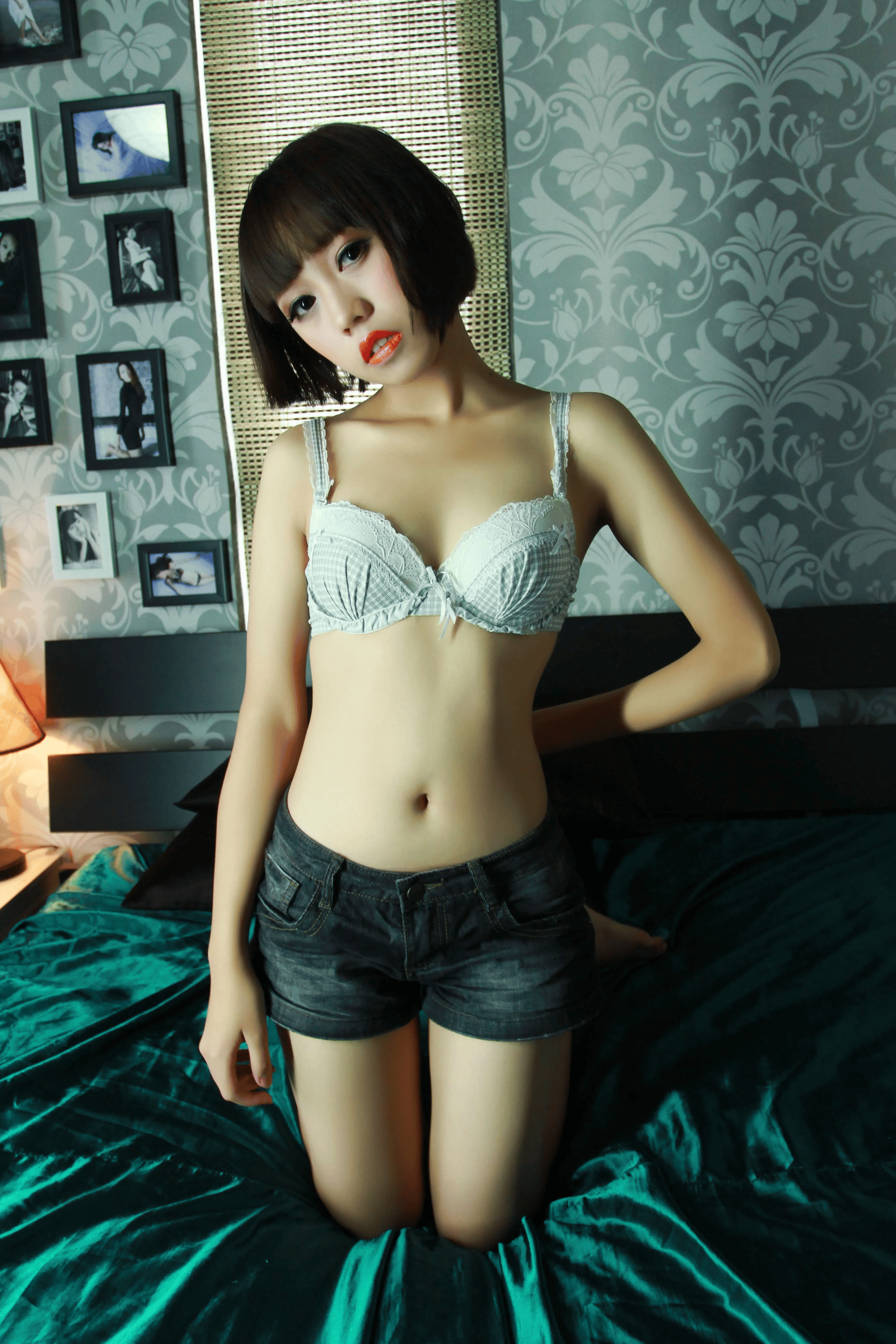 Young East Asian woman with short dark bob cut and bangs, kneeling on a bed covered with dark teal satin bedding. She is wearing a light gingham lace-trimmed bra and dark-wash denim shorts, looking toward the camera. The background features damask patterned wallpaper mounted with framed photos, and a bamboo window blind behind her.