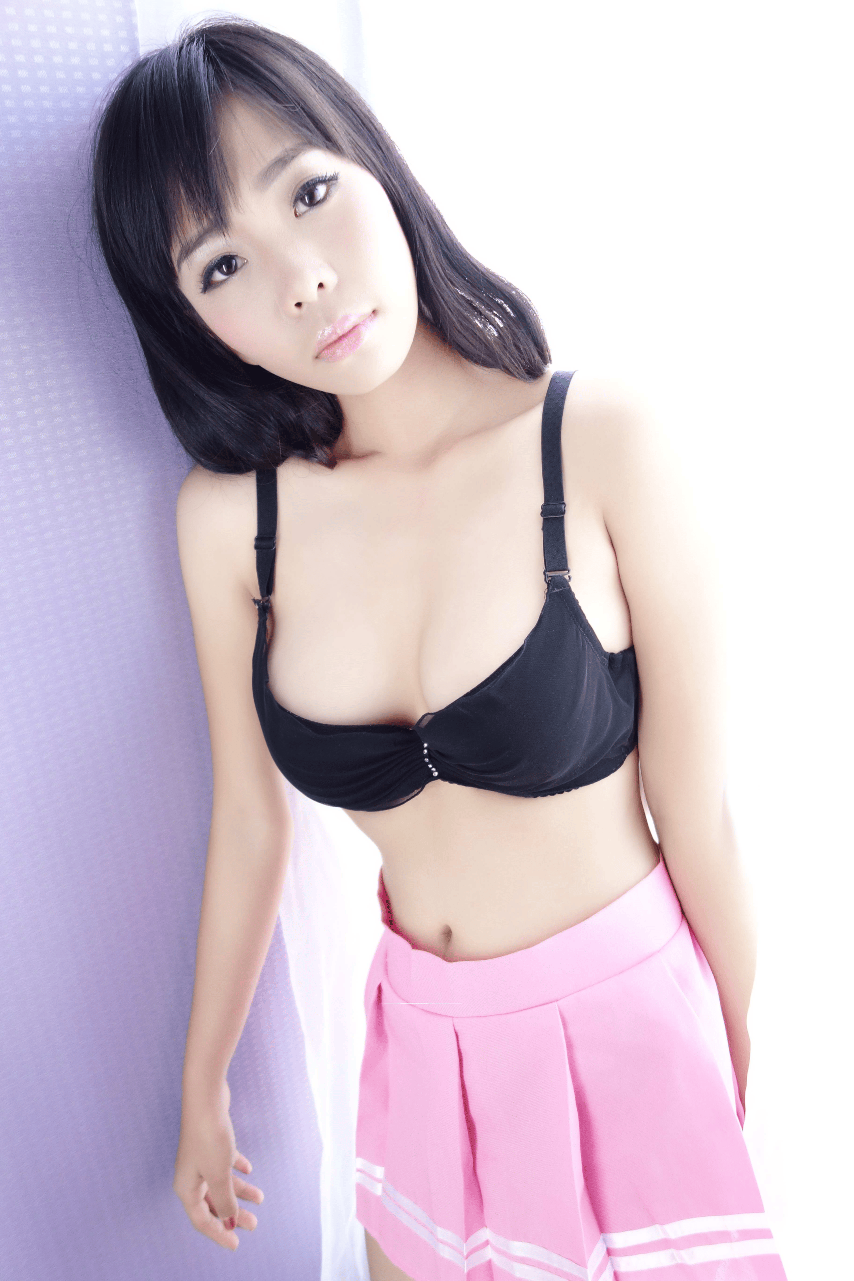 Young East Asian woman with shoulder-length dark hair and bangs, wearing a black bra with small rhinestone accents at the center and a pink pleated mini skirt with white stripe details, leaning against a light purple textured curtain, looking directly at the camera in a bright indoor setting.